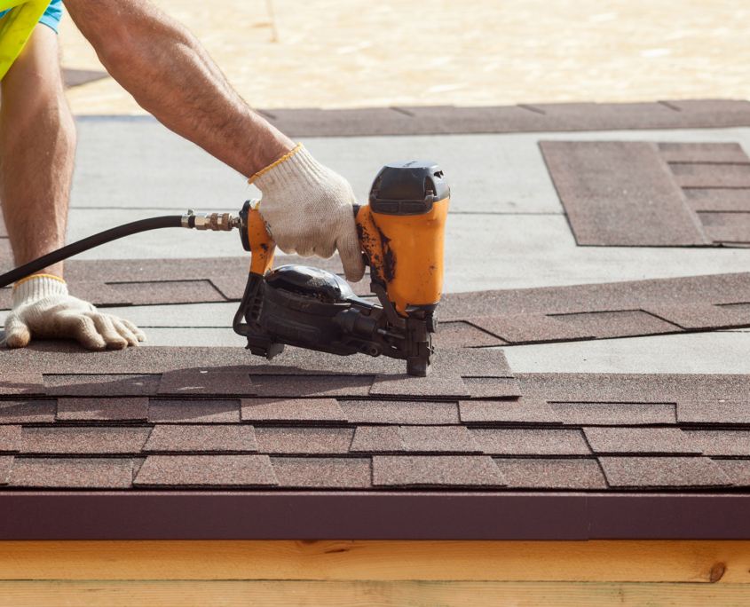 Construction Worker Putting The Asphalt Roofing (shingles) With Nail Gun On A New Frame House